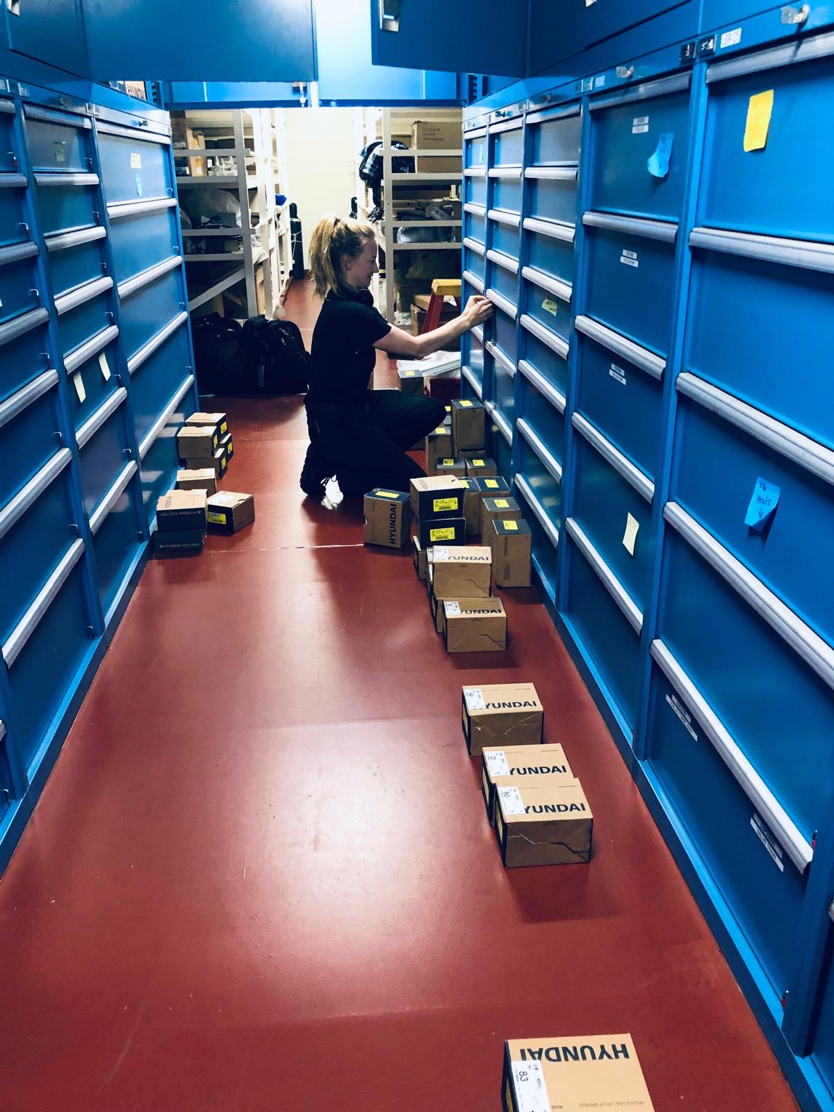 Never-Lo's founder kneeling in a shipboard storeroom aisle, labeling and verifying blue parts drawers while boxed Hyundai spares are staged on the deck for sorting.