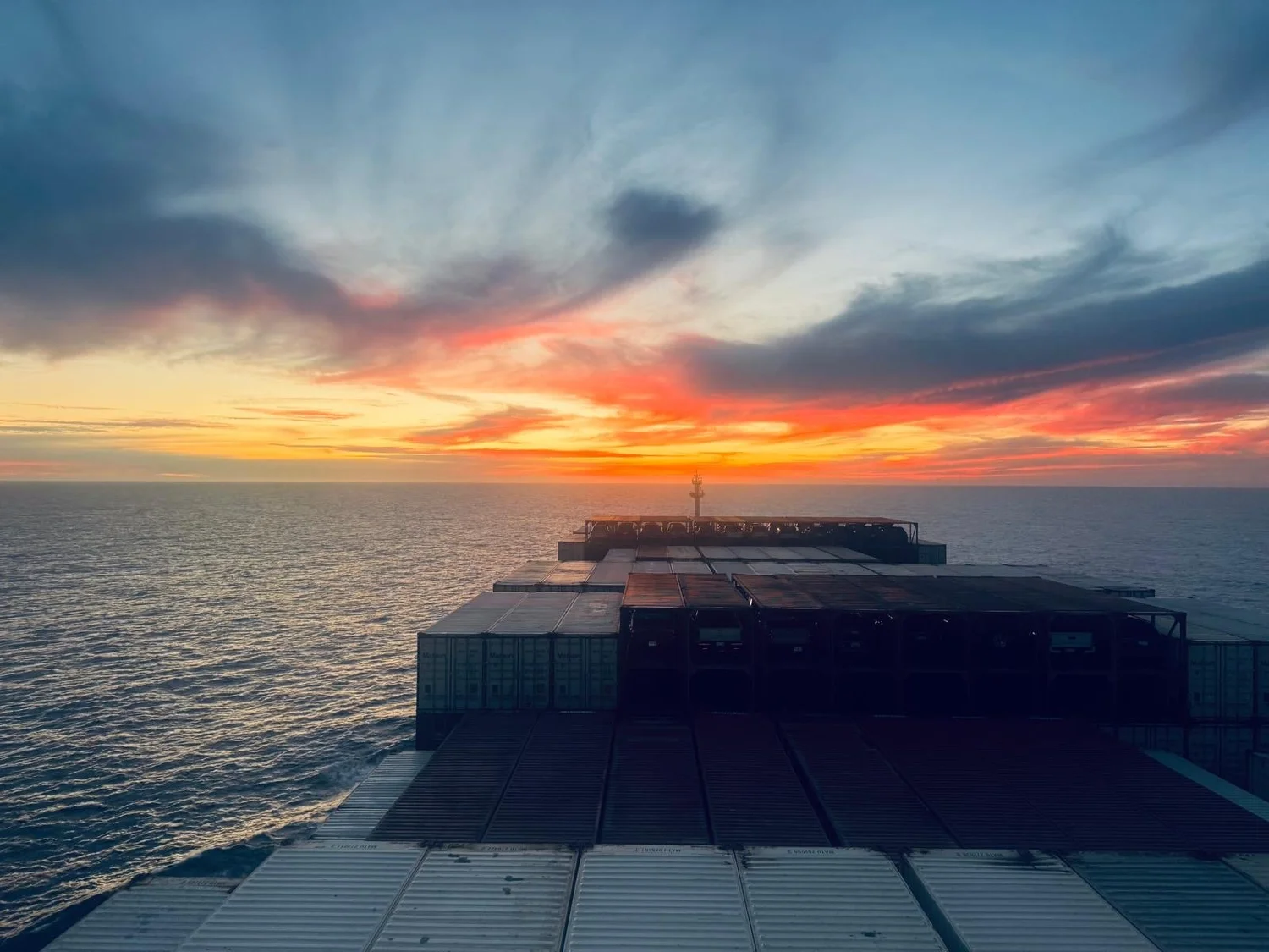View forward over a containership's stacked deck cargo at sunset, open ocean ahead on both sides.