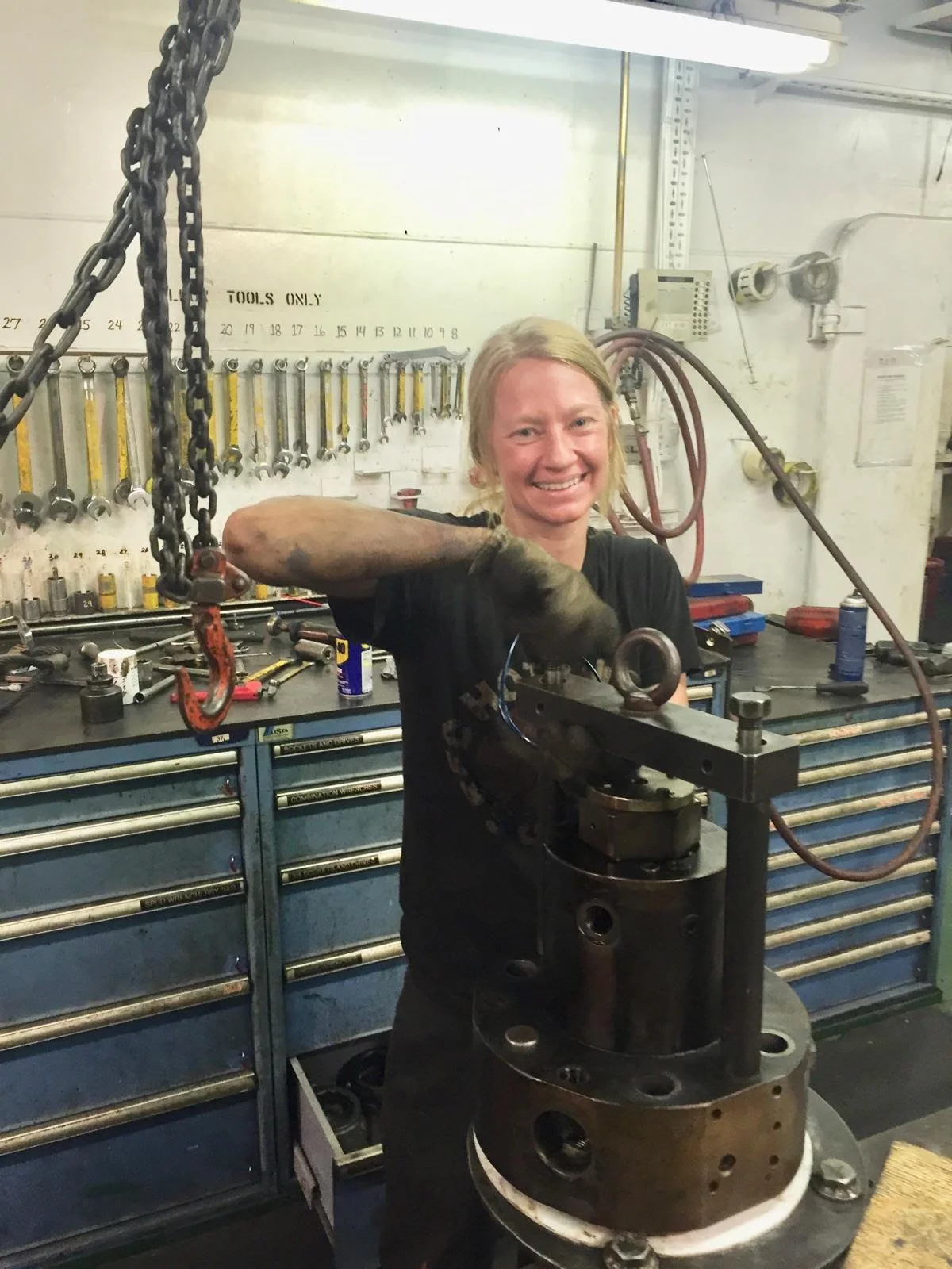 Never-Lo's founder in a ship's engine workshop, hand on a cylinder head assembly rigged to a chain hoist, with a wrench board and tool cabinets behind her.