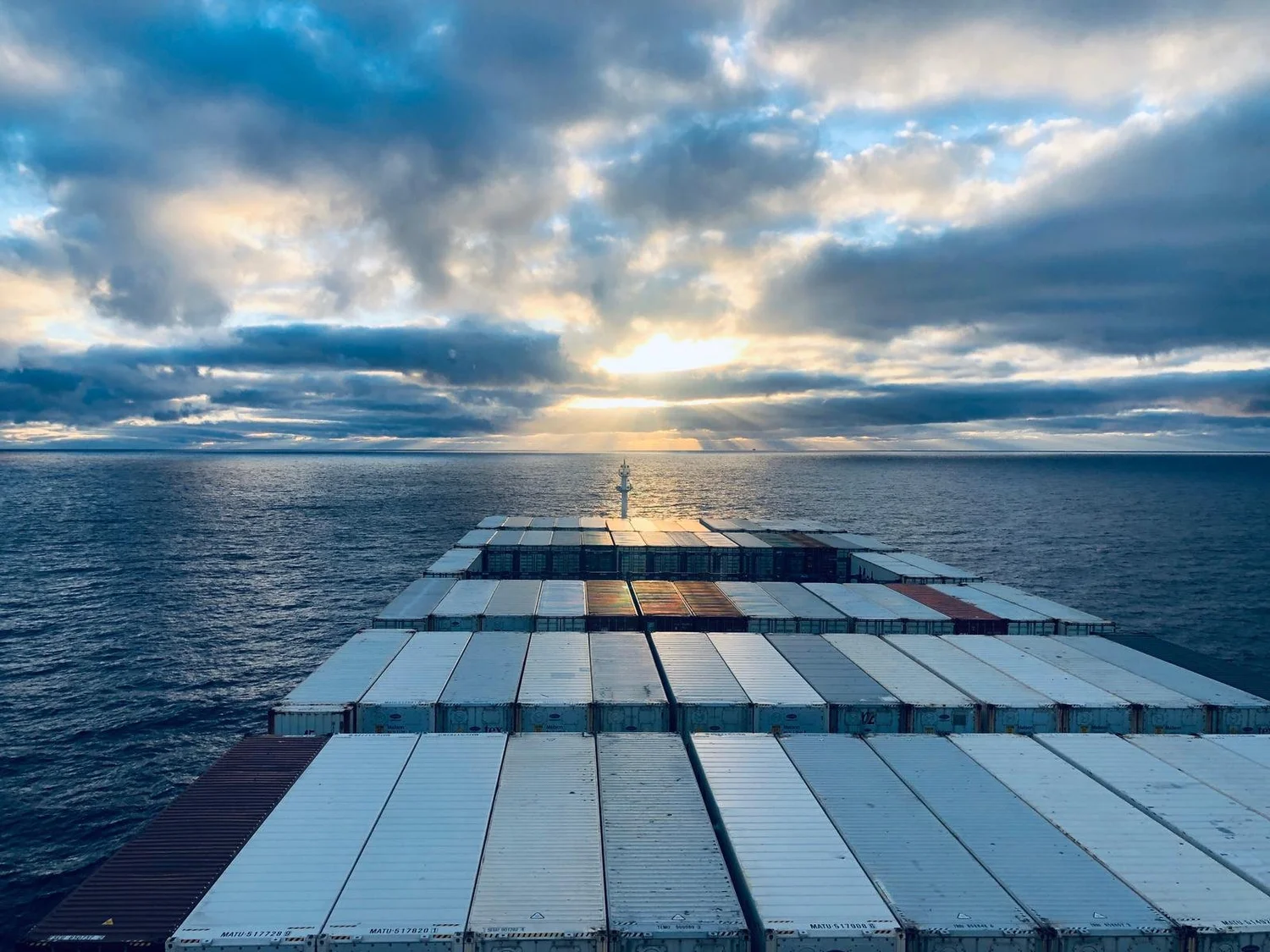 A loaded containership underway at sea, bow wave breaking along the hull.